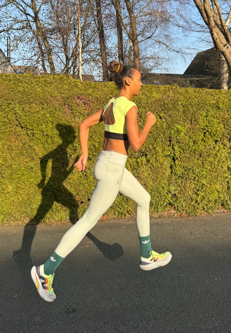 Νεαρή γυναίκα τρέχει σε εξωτερικό χώρο σε ασφαλτοστρωμένο μονοπάτι, φορώντας ανοιχτό κίτρινο κοντό τοπ, ανοιχτόχρωμο κολάν, πράσινες κάλτσες και νέον αθλητικά παπούτσια κατά τη διάρκεια της ημέρας.