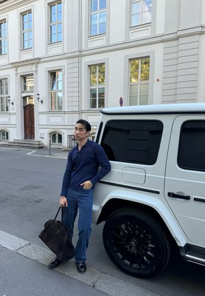 Bolso tote de ante negro, camisa de manga larga azul marino, jeans de color azul claro y zapatos oscuros al lado de un SUV blanco con ruedas negras contra un fondo de calle.