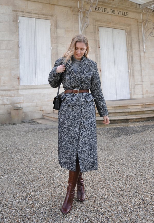 Texturierter schwarz-weißer Wollmantel mit tailliertem Gürtel, tiefem V-Ausschnitt und Knöpfen vorne, kombiniert mit braunen kniehohen Stiefeln.