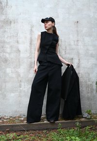 Top nero senza maniche con orlo asimmetrico, abbinato a pantaloni neri a gamba larga. Un blazer nero abbinato è tenuto nella mano destra.