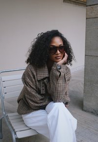 Cappotto oversize marrone a motivo pied de poule, pantaloni bianchi, grandi occhiali da sole scuri, seduto su una sedia contro una parete neutra con pietra testurizzata.