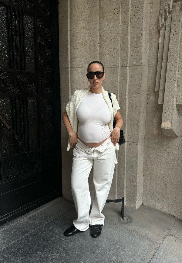 Femme vêtue de vêtements blancs et de lunettes de soleil, debout contre un mur en pierre à côté d'une porte métallique décorative noire.
