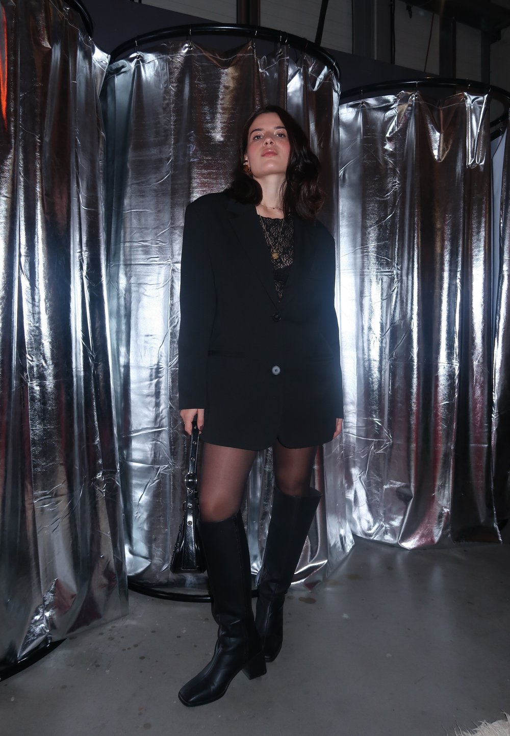 Μαύρο υπερμεγέθες σακάκι πάνω από μια διαφανή δαντελένια μπλούζα, συνδυασμένο με μαύρες δερμάτινες μπότες μέχρι το μηρό και κρατώντας μια μικρή μαύρη τσάντα.