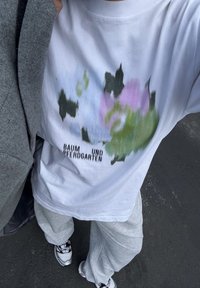 White cotton T-shirt featuring an abstract floral design in green and pink, with black text reading "BAUM UND PFERDGARTEN." Gray pants and sporty shoes visible.
