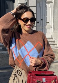 Bruine oversized trui met een blauw en oranje ruitpatroon, gecombineerd met een geruite rok en een rode, tekstuurlijke handtas. Bruine zonnebril maakt de look compleet.