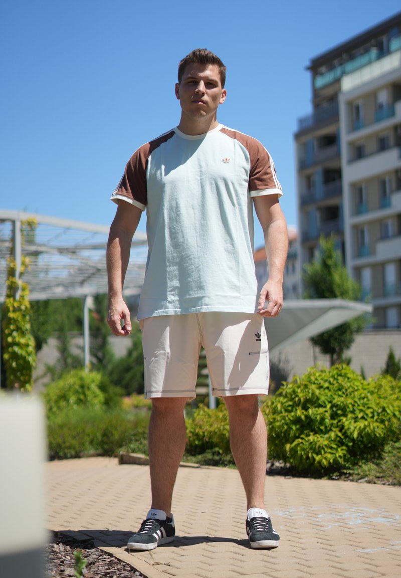 Junger Mann steht draussen und trägt ein hellblaues und braunes kurzärmeliges Hemd, beige Shorts und schwarze Turnschuhe an einem sonnigen Tag.