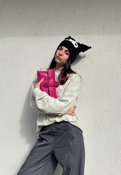 Joven con un gorro de panda y un suéter ligero sostiene un bolso de mano rosa brillante, de pie frente a una pared texturizada de color gris claro.