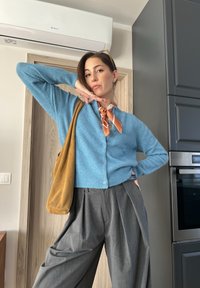 Ανοιχτό μπλε μάλλινο cardigan με V-λαιμόκοψη και κουμπιά, συνδυασμένο με γκρι κομμένα παντελόνια με πτυχές. Ένα καφέ σουέτ τσάντα ώμου και ένα patterned κασκόλ συμπληρώνουν την εμφάνιση.