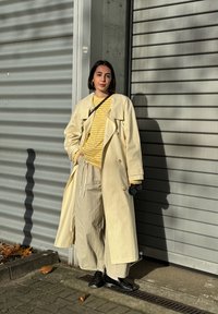 Beige trench coat over a yellow striped shirt with light beige trousers. Black shoes and a camera. Shadow patterns on a textured wall.