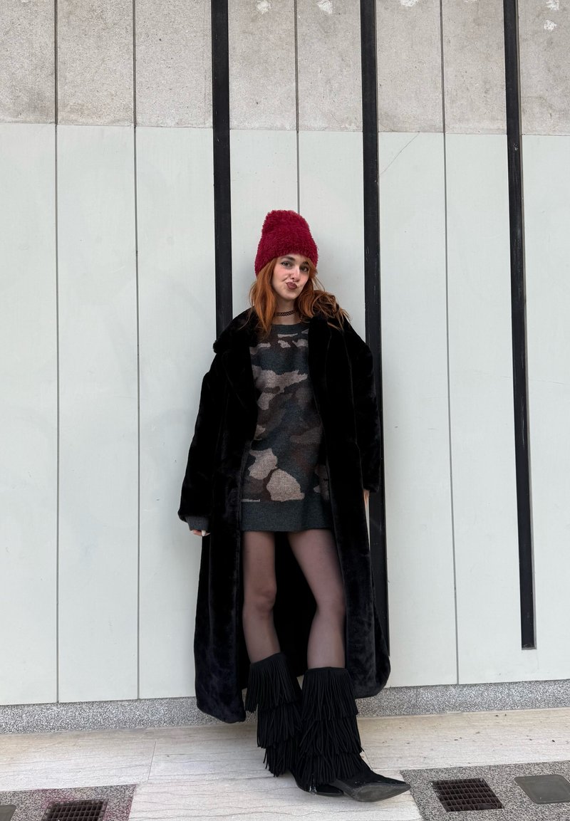 Manteau en fausse fourrure noire, robe-pull à motif camouflage, bottes noires à franges et bonnet en tricot rouge devant un mur abstrait gris.