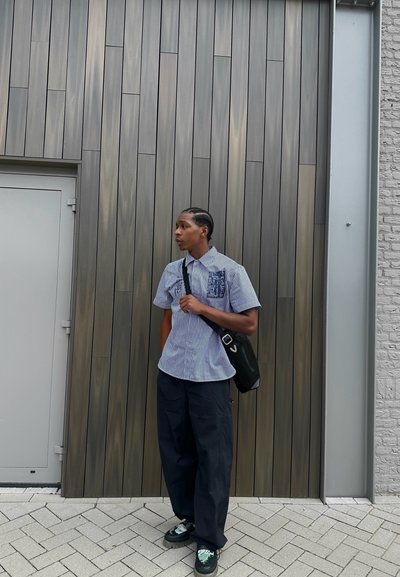 Camisa de manga corta a rayas azul con bolsillo, pantalones oscuros holgados y bolso negro; apoyado contra una pared de madera y piedra texturizada.