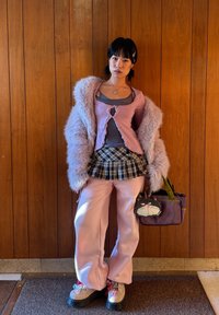 Jeune femme portant des vêtements superposés rose et gris avec une jupe à carreaux et un manteau duveteux, tenant un sac violet avec un visage de chat devant un mur en panneaux de bois.