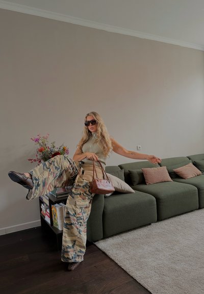 Mujer con cabello largo y rubio, vestida con pantalones de camuflaje, top sin mangas, gafas de sol y sosteniendo un bolso marrón, sentada en un sofá verde oscuro.