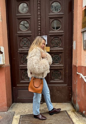 Mujer vestida con una chaqueta beige de felpa, jeans azul claro y botas marrones, de pie frente a una puerta marrón sosteniendo un bolso marrón.