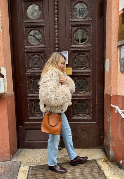 Mujer vestida con una chaqueta beige de felpa, jeans azul claro y botas marrones, de pie frente a una puerta marrón sosteniendo un bolso marrón.
