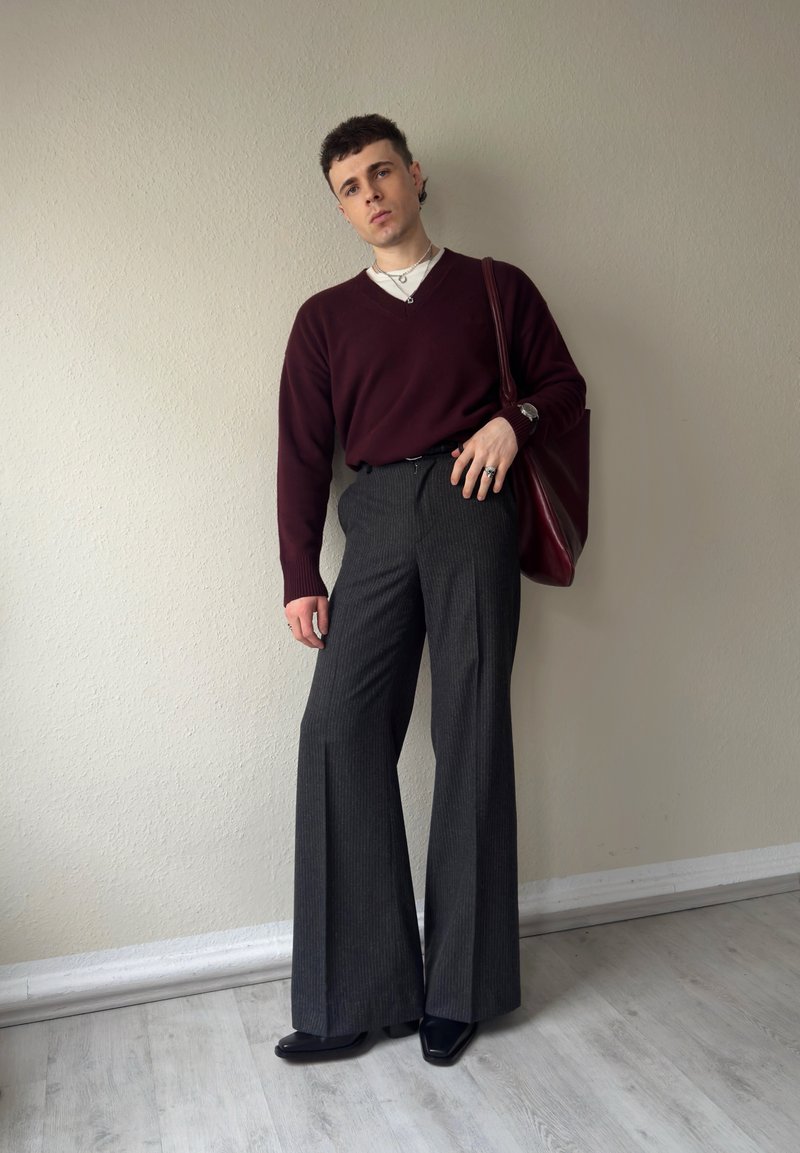 Junge Person trägt einen kastanienroten Pullover, graue weite Hosen, schwarze Schuhe und hält eine passende kastanienrote Tragetasche vor einer schlichten Wand.