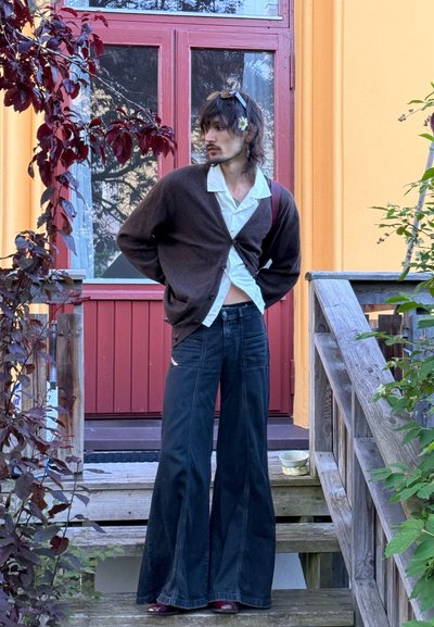 Cárdigan de punto marrón sobre una camisa blanca con botones, combinado con pantalones amplios de mezclilla negra. De pie en unas escaleras de madera, rodeado de plantas.