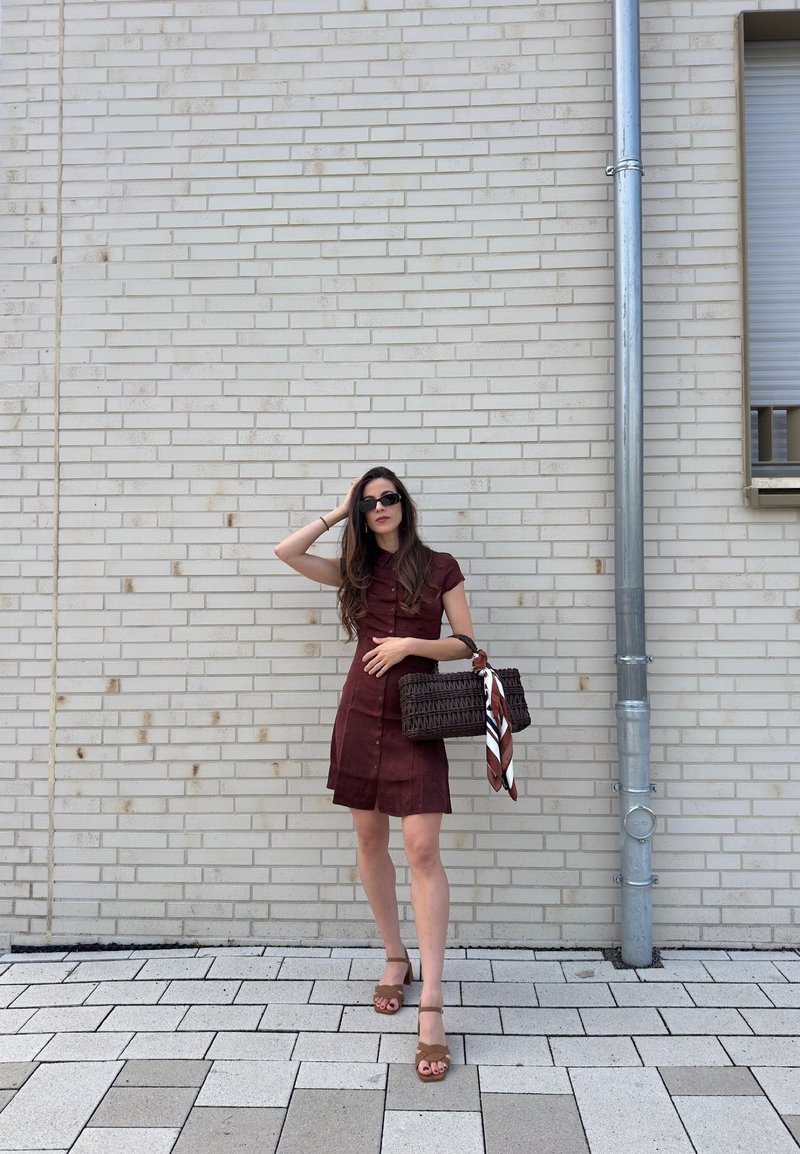 Donna in un vestito marrone a maniche corte e sandali che tiene una borsa intrecciata con una sciarpa, in piedi contro un muro di mattoni chiari accanto a una grondaia metallica.