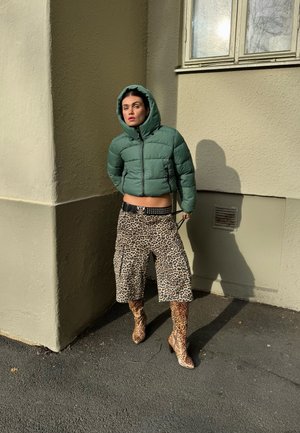 Chaqueta acolchada verde con capucha, pantalones cortos cargo con estampado de leopardo y botas de tacón a juego con el mismo estampado. La textura es suave y con un aspecto brillante.