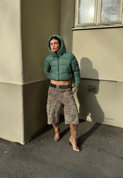 Chaqueta acolchada verde con capucha, pantalones cortos cargo con estampado de leopardo y botas de tacón a juego con el mismo estampado. La textura es suave y con un aspecto brillante.