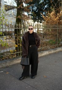 Cappotto in pelliccia sintetica marrone, dolcevita nero, pantaloni neri larghi, scarpe nere robuste e una borsa nera, sullo sfondo di una recinzione texturizzata e verdure.