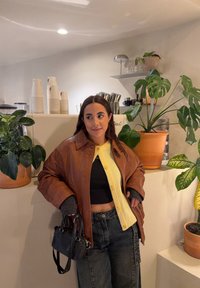 Femme en veste en cuir marron et cardigan jaune debout près du comptoir à café avec des plantes en pot, tenant un petit sac à main noir.