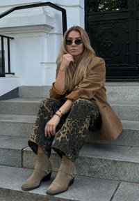 Καφέ σουέτ oversized σακάκι, παντελόνι σε ζωντανό σχέδιο με φαρδιά πόδια και ταμπά μπότες μέχρι τον αστράγαλο με καρφίτσες, καθισμένες σε πέτρινες σκάλες.