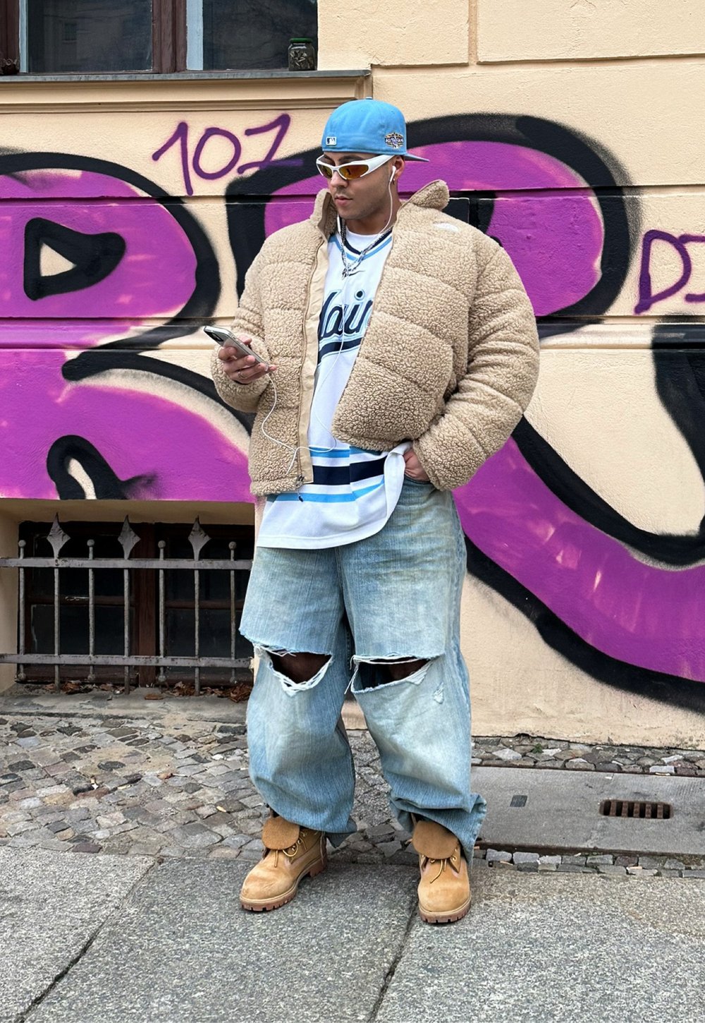 Μπεζ τζάκετ από σέρπα, λευκό μπλουζάκι με μπλε λεπτομέρειες, φθαρμένο ανοιχτό μπλε τζιν, καφέ μπότες και γυαλιά ηλίου. Αστικό φόντο με γκράφιτι.