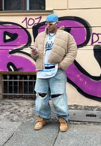 Μπεζ τζάκετ από σέρπα, λευκό μπλουζάκι με μπλε λεπτομέρειες, φθαρμένο ανοιχτό μπλε τζιν, καφέ μπότες και γυαλιά ηλίου. Αστικό φόντο με γκράφιτι.