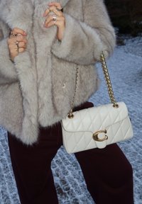 Άτομο που φορά μια αφράτη μπεζ γούνα και μπορντό παντελόνι, κρατώντας μια λευκή τσάντα χειρός με καπιτονέ υφή και χρυσή αλυσίδα-λαβή και κλείσιμο.