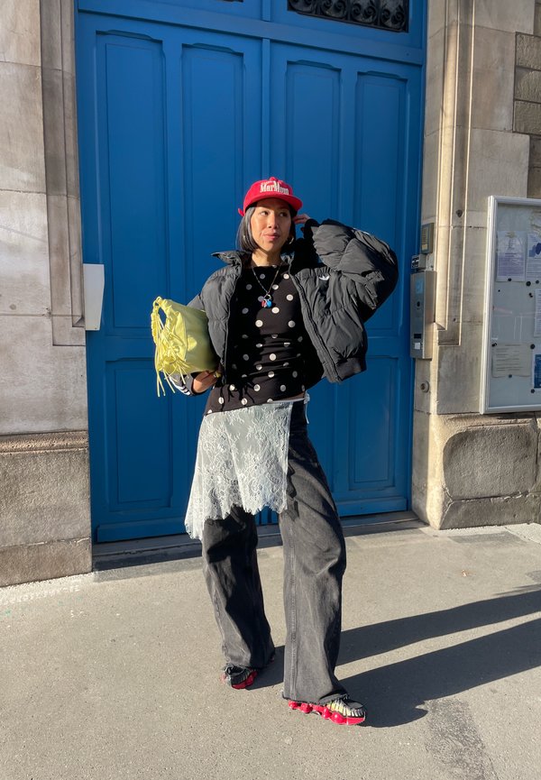 Sieviete melnā pūkainā jaku, melnu topu ar polka punktiem, platām džinsa biksēm, sarkanu cepuri, tur dzeltenu somu, stāvot pie lielām zilām durvīm.