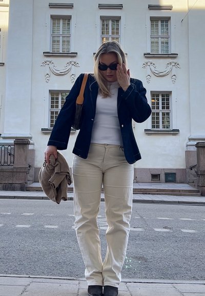 Blazer azul marino sobre una blusa blanca, pantalones crema y botas negras. Sosteniendo un suéter color camello y un bolso de hombro marrón. Se observa un entorno urbano.