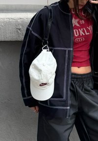 Personne portant une veste noire avec des coutures blanches, un top rouge raccourci "Brooklyn USA", tenant une sangle noire avec une casquette Nike blanche attachée.