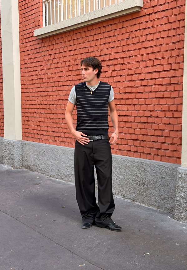 Un joven con un chaleco de punto a rayas y unos pantalones negros holgados está de pie en la acera junto a una pared de ladrillo rojo y mira hacia un lado.