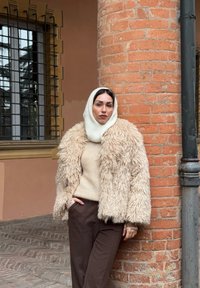 Femme portant une capuche tricotée beige et une veste crème duveteuse, appuyée contre une colonne en briques rouges près d'une fenêtre avec des barreaux noirs.