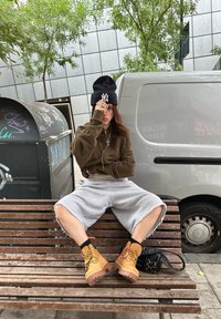 Sweat à capuche marron, short gris coupé, bottes jaunes avec des accents rouges, bonnet noir avec logo, collier argenté, assis sur un banc en bois près d'une camionnette.