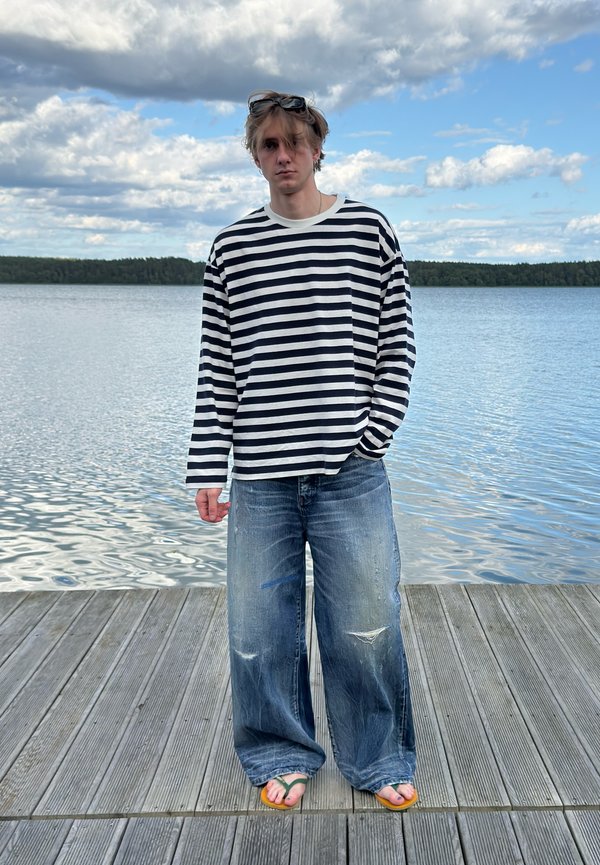 Junge Person, die ein gestreiftes langärmeliges Shirt, weite Jeans und Sandalen trägt, steht auf einem Holzsteg an einem ruhigen See unter einem bewölkten Himmel.