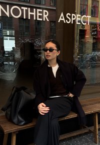 Manteau noir oversize, haut blanc, cardigan marron et pantalon à fines rayures. Sac à main noir et lunettes de soleil. Banc en bois dans un cadre urbain.