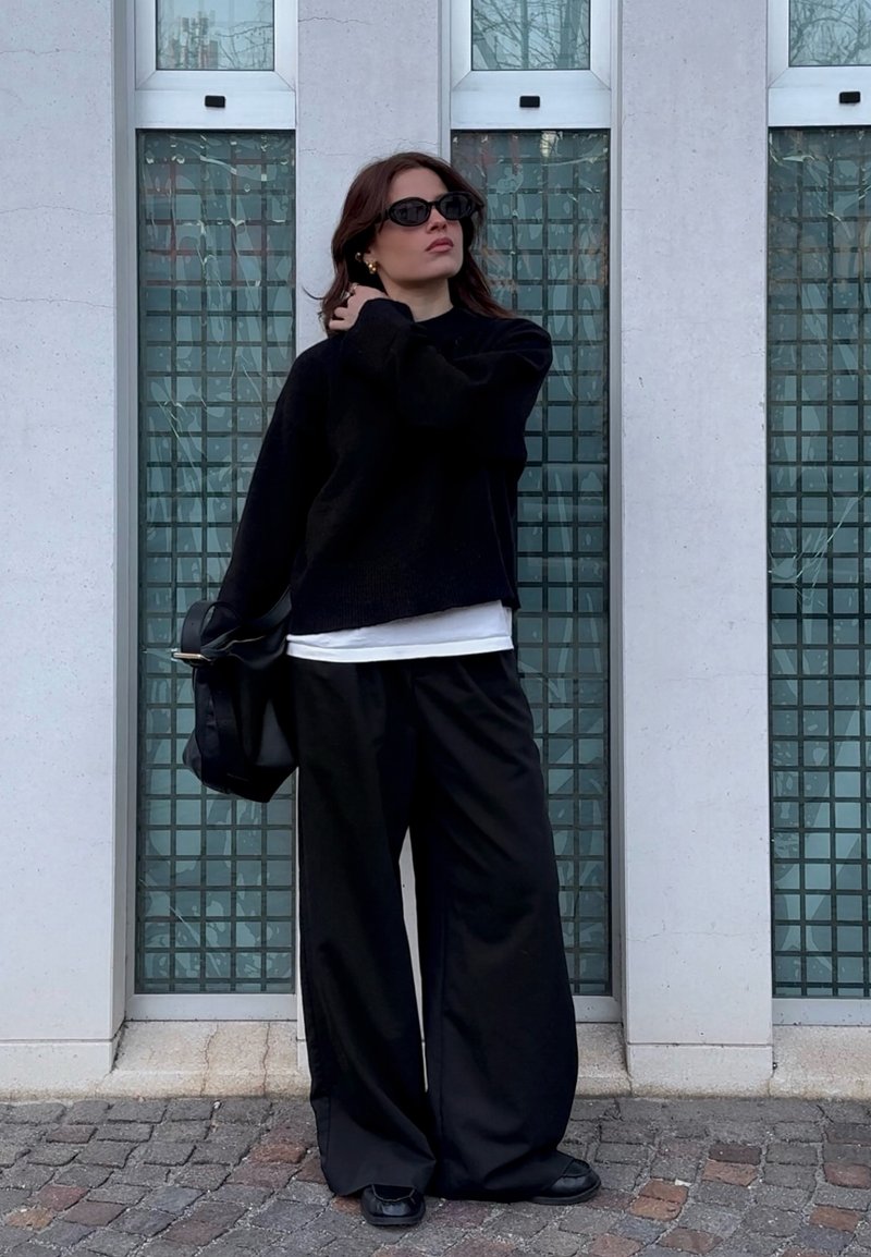 Donna con occhiali da sole neri, maglione nero, pantaloni neri larghi, che tiene una borsa nera, in piedi contro un muro bianco con finestre a griglia.