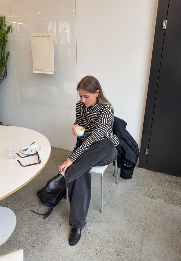 Frau mit gestreiftem Shirt und schwarzer Hose sitzt im Schneidersitz auf einem Stuhl und hält eine Tasse Kaffee neben einem Tisch mit Sonnenbrille, Glas und Teller.