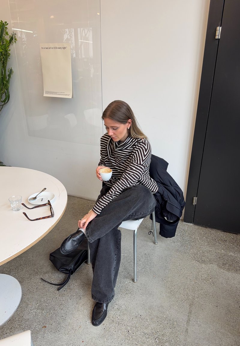 Woman in striped shirt and black pants sits cross-legged on chair, holding a cup of coffee next to table with sunglasses, glass, and plate.