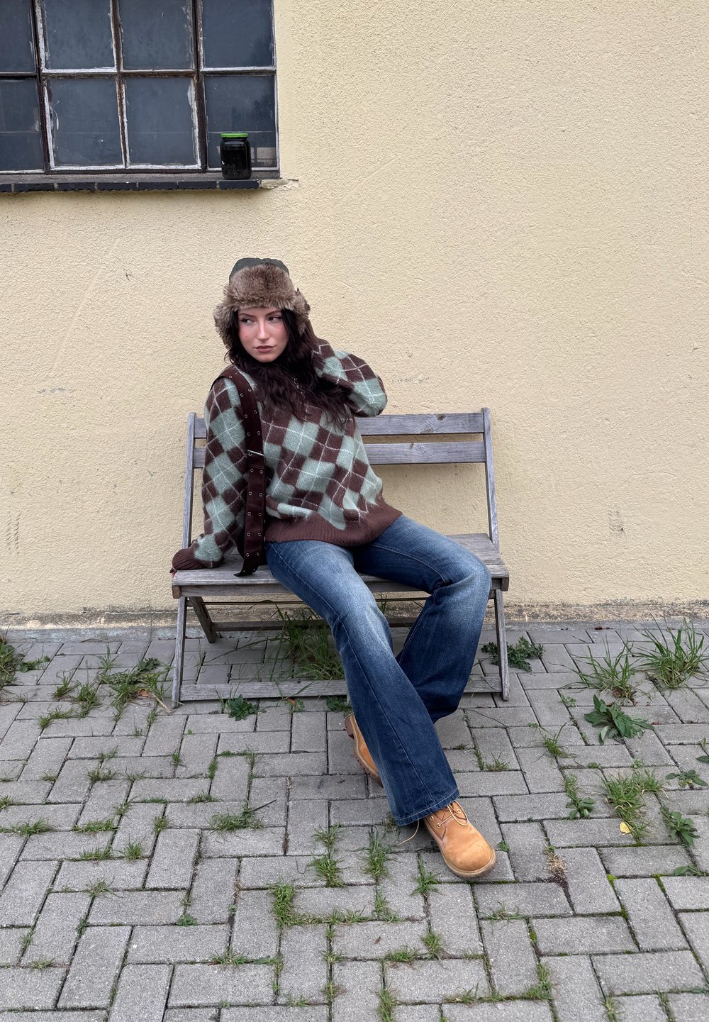 Brown and green argyle sweater, flared denim jeans, tan boots, and a fur hat. Sitting on a wooden bench against a textured yellow wall.