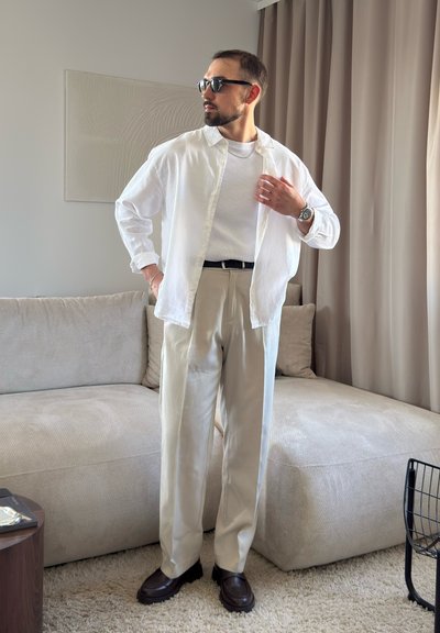 Hombre que lleva camisa blanca, camiseta blanca, pantalones beige, cinturón negro y zapatos marrones, de pie en una sala de estar con sofá beige y cortinas.