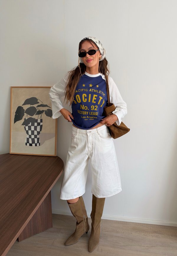 Femme portant des lunettes de soleil, un foulard blanc, une chemise bleue à manches longues, un short blanc jusqu'aux genoux et des bottes marron à côté d'une image encadrée de plante.