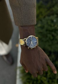 Χρυσό ρολόι με μπλε καντράν, ρωμαϊκούς αριθμούς, παράθυρο ημερομηνίας και χαρακτηριστικά χρονογράφου, συνδεδεμένο με μια υφασμένη χρυσή βραχιόλι.