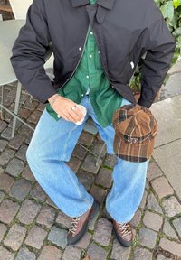 Giacca bomber nera sopra una camicia a righe verdi, jeans a gamba larga azzurri, scarpe da trekking marroni e un berretto a quadri marrone posato sulle ginocchia.