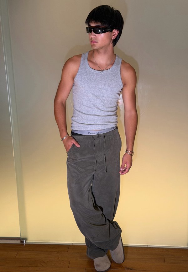 Jeune homme portant des lunettes de soleil, un débardeur gris, un pantalon ample foncé et des bracelets, debout avec une main dans la poche contre un mur beige uni.
