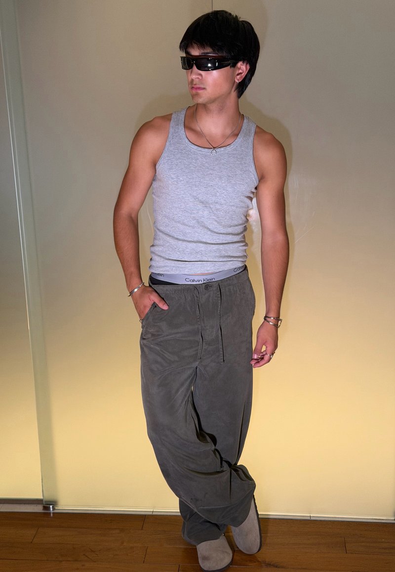 Jeune homme portant des lunettes de soleil, un débardeur gris, un pantalon ample foncé et des bracelets, debout avec une main dans la poche contre un mur beige uni.