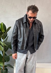 Veste en cuir noir avec fermeture éclair à l'avant, grand col et deux poches latérales. Associée à une chemise noire à boutons et un pantalon gris clair.