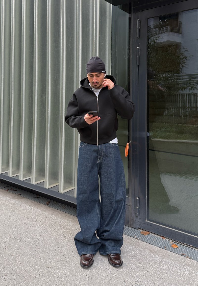 Homme portant un sweat à capuche noir, un bonnet, un jean large et des chaussures marron, debout à l'extérieur près d'une porte en verre, regardant son téléphone et touchant son écouteur.